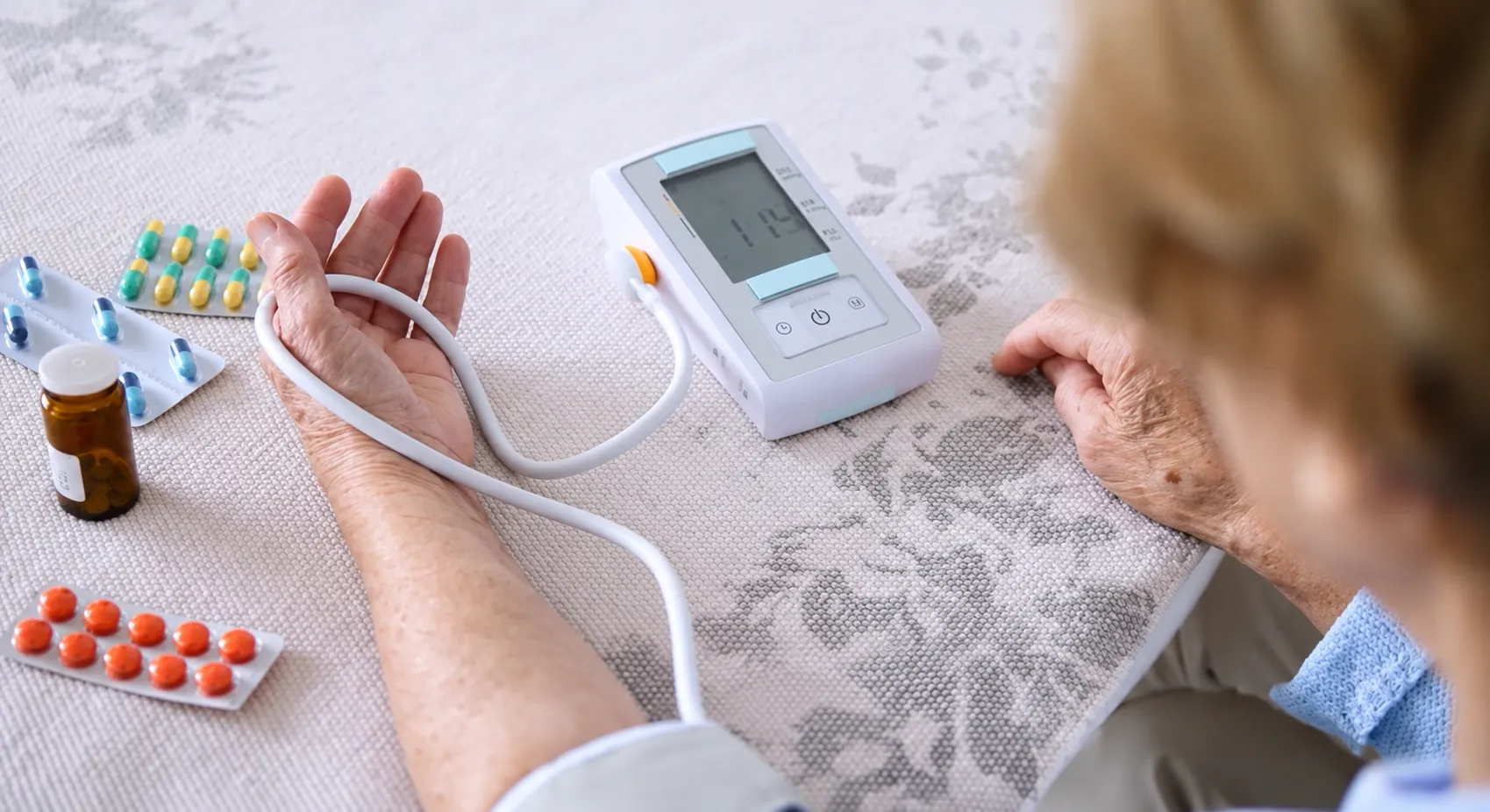 An elderly man holding a handful of blood pressure pills beside a monitor showing an elevated reading